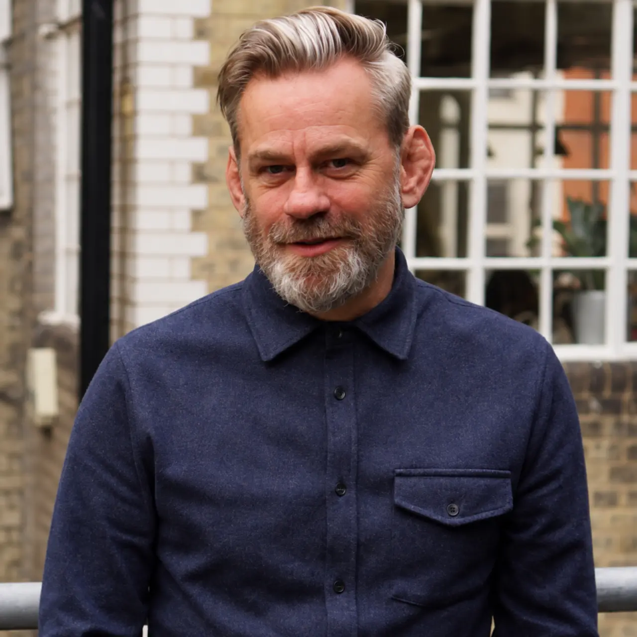 Niall Murdoch, a man with grey hair and beard wearing a dark navy shirt, smiles at the camera in front of a brick building with white-framed windows