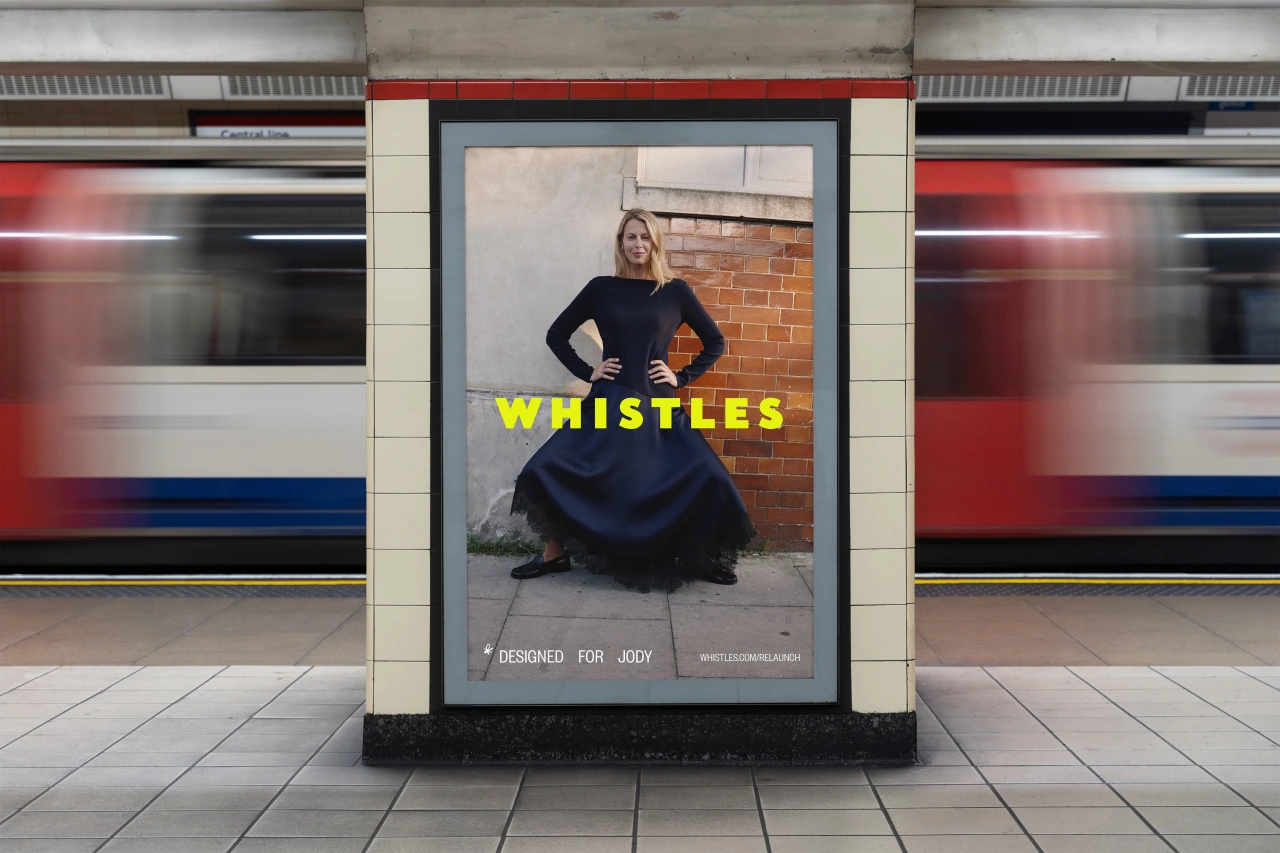 Woman in black dress standing against brick wall on London Underground platform with Whistles advertisement displaying yellow "WHISTLES" text
