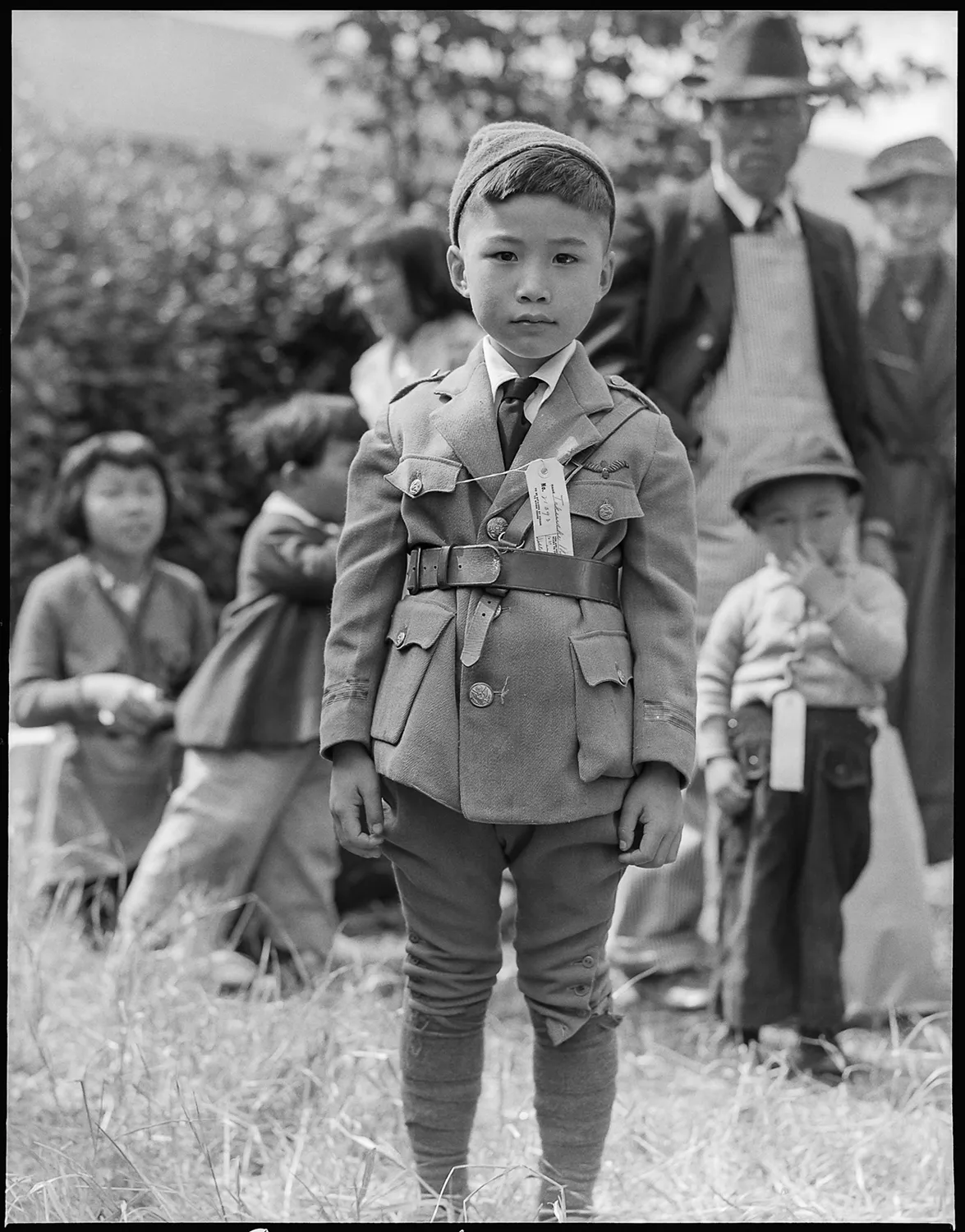 Young boy in military-style uniform with cap and belt standing in front of gathered adults and children at Manzanar internment camp