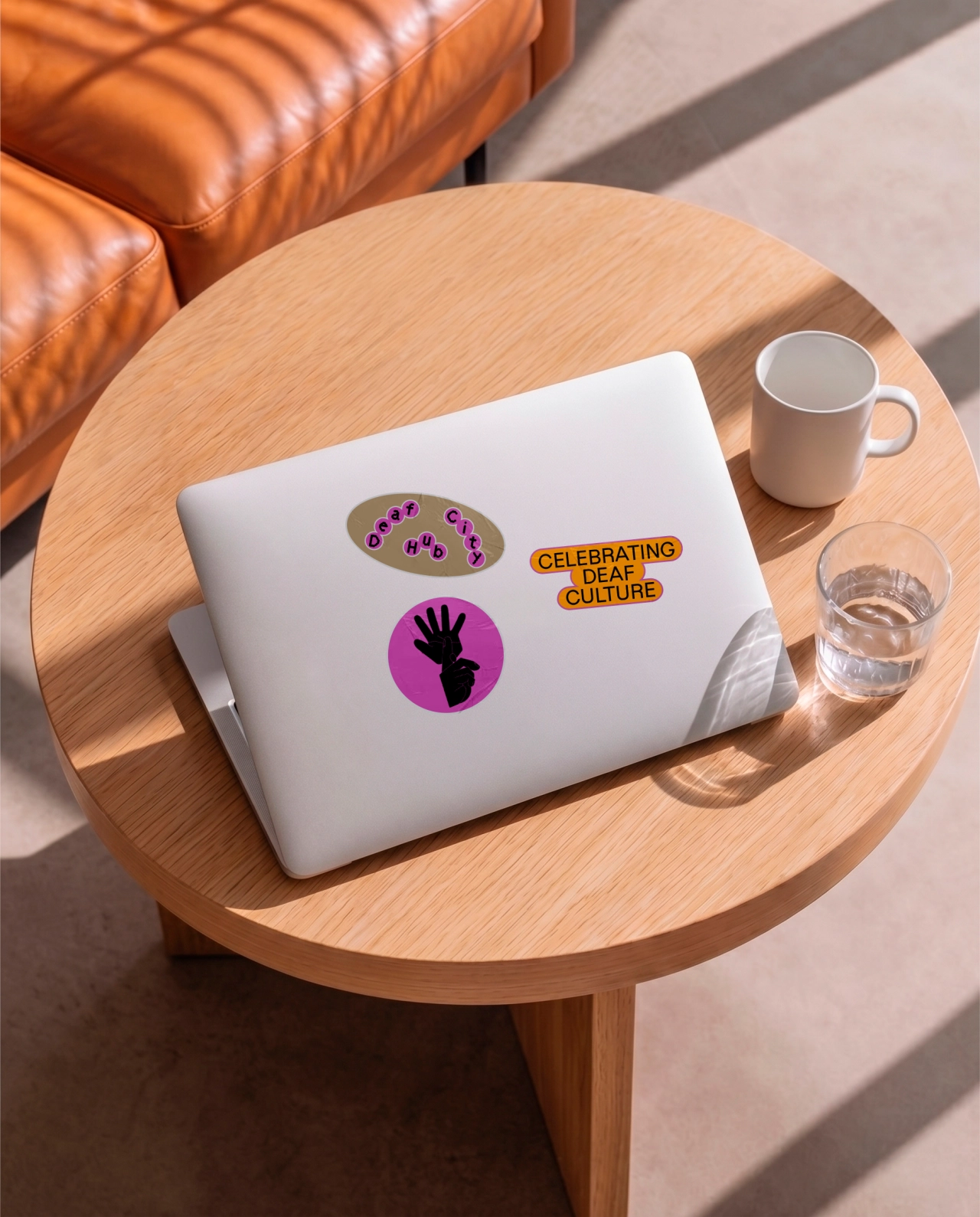White laptop with DeafCity Hub stickers including purple circles representing hand gestures from British Sign Language, on a wooden table with coffee mug and water glass