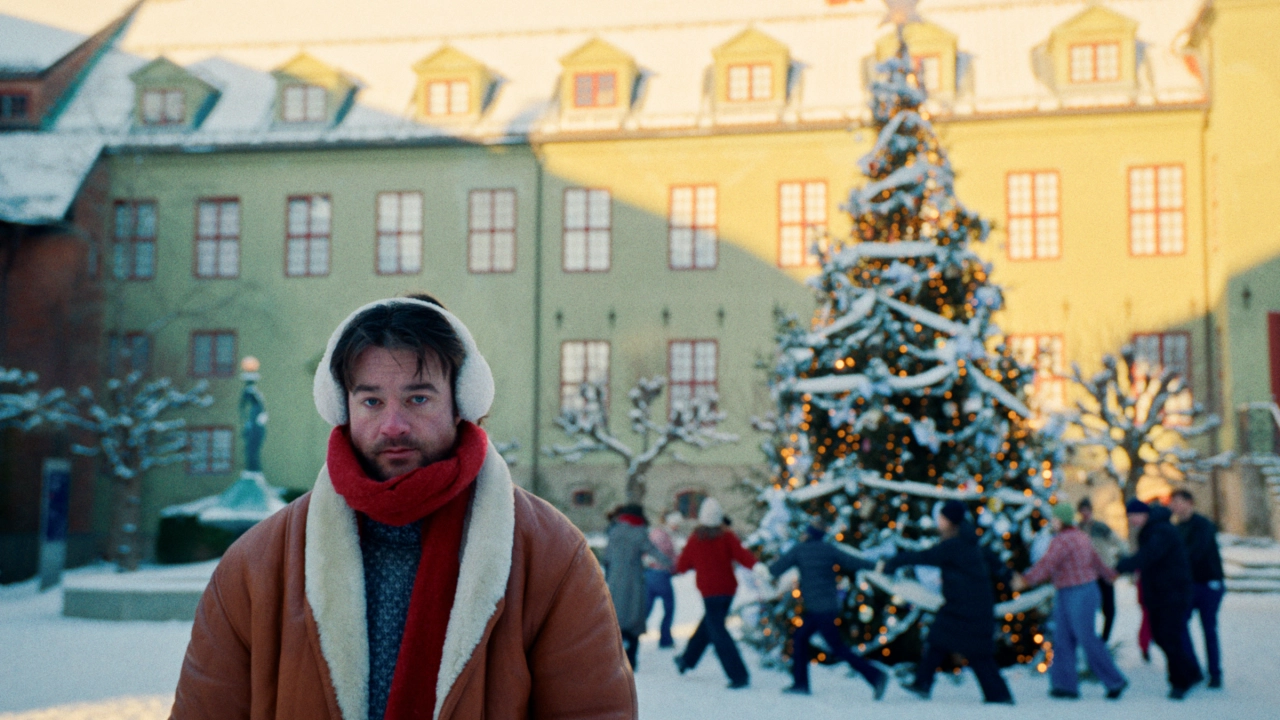 Halfdan stands in a snowy Oslo city square wearing a brown coat and red scarf, with a decorated Christmas tree and colourful historic buildings behind him
