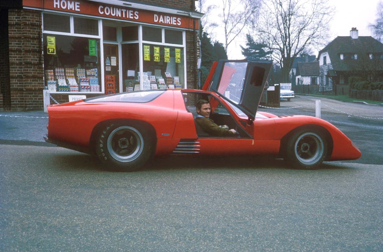 Red McLaren M6GT sports car with gullwing door open, parked outside a vintage service station with "Home Counties Dairies" signage
