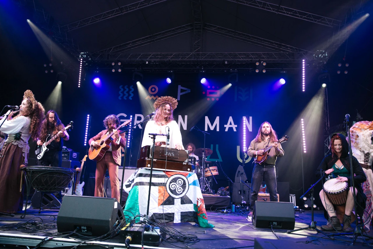 A folk music ensemble with traditional instruments and headpieces performs live on a stage lit by blue spotlights.