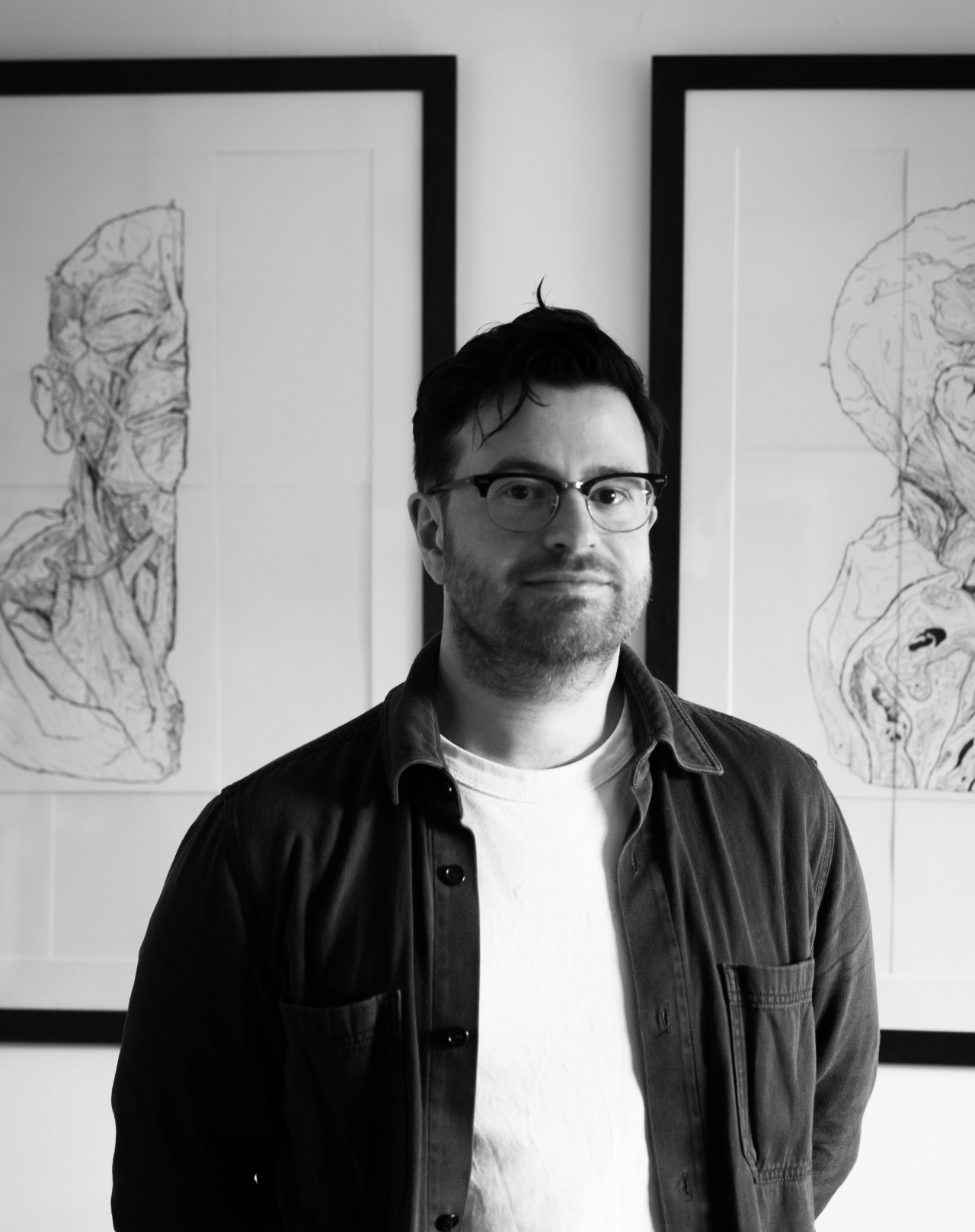 Man wearing glasses and dark shirt standing in front of two framed ink drawings of faces on a white wall
