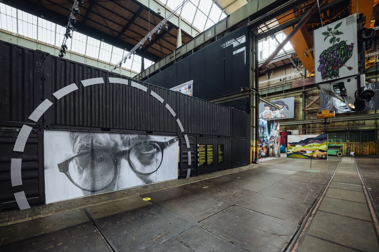 Large-scale black and white mural of a close-up face wearing glasses inside an industrial warehouse gallery space with yellow overhead cranes