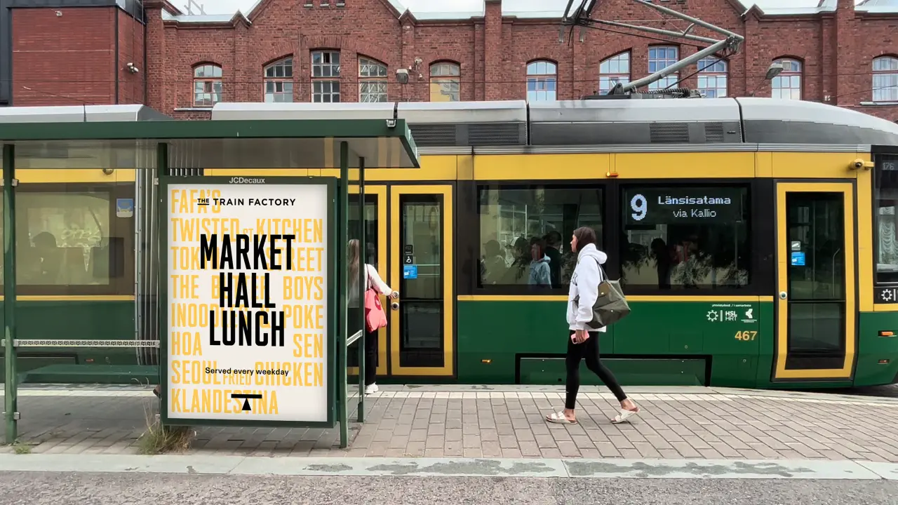 A yellow and green tram branded with The Train Factory's number 9 route stops outside a brick building, with a Market Hall Lunch poster visible on the shelter beside it.