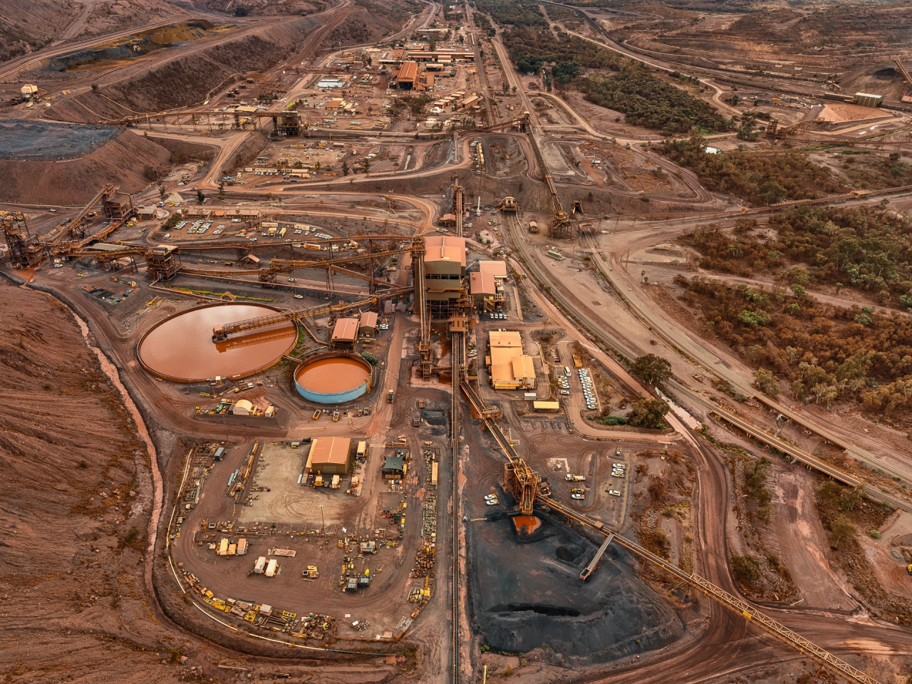 Aerial view of a large mining operation with orange processing tanks, dark water ponds, industrial buildings and excavation equipment spread across a barren landscape with roads and cleared areas