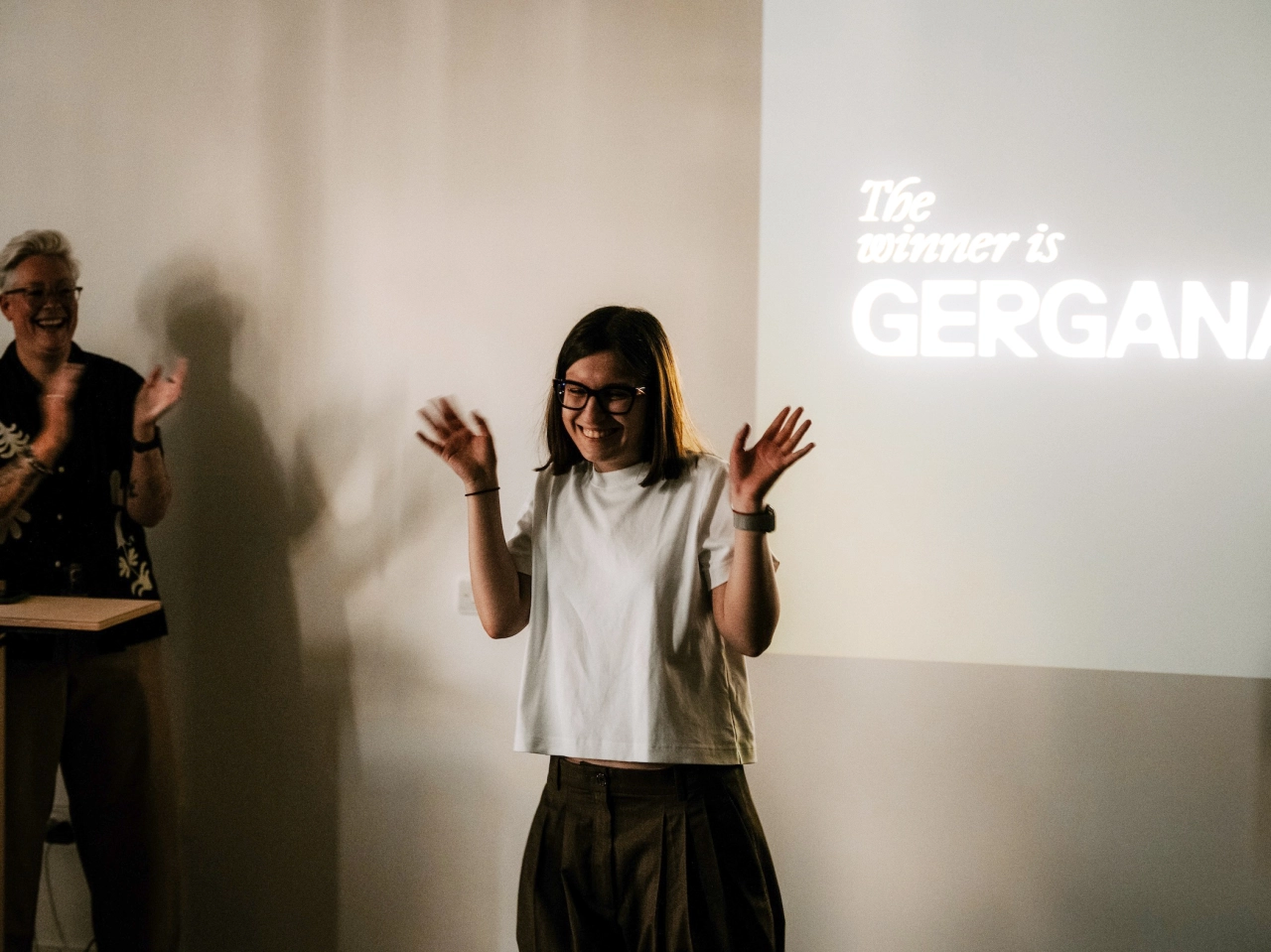 Person wearing glasses and white t-shirt gesturing expressively in front of projected text reading "The winner is GERGANA" at what appears to be an awards event