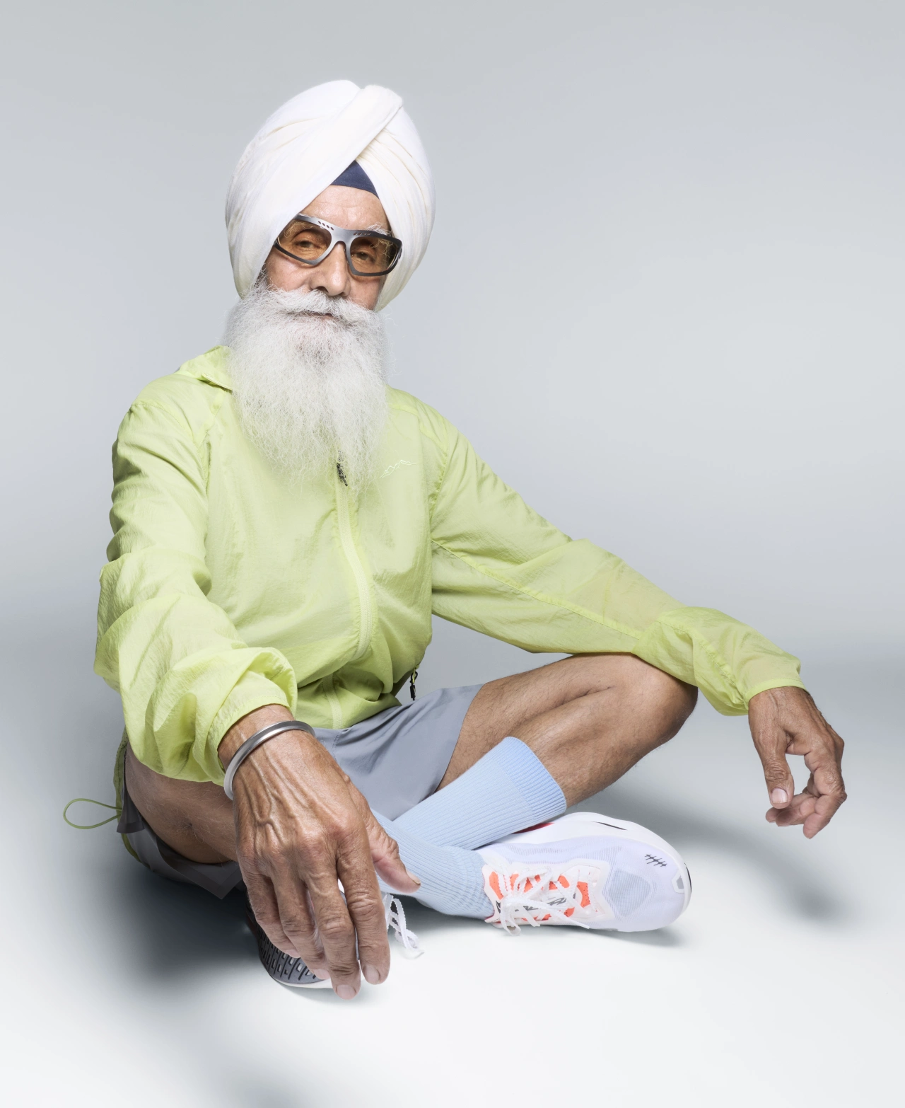 Man in white turban and glasses wearing bright yellow jacket and white socks, seated against grey background as part of Altra's Legacy Team campaign photographed by Rankin