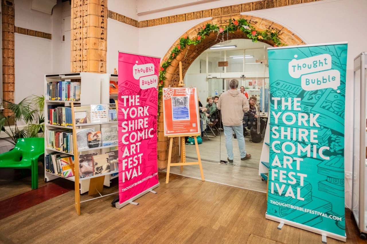 Colourful pink and turquoise banners for The Yorkshire Comic Art Festival stand in a venue's entrance hall with visitors and a bookshelf visible