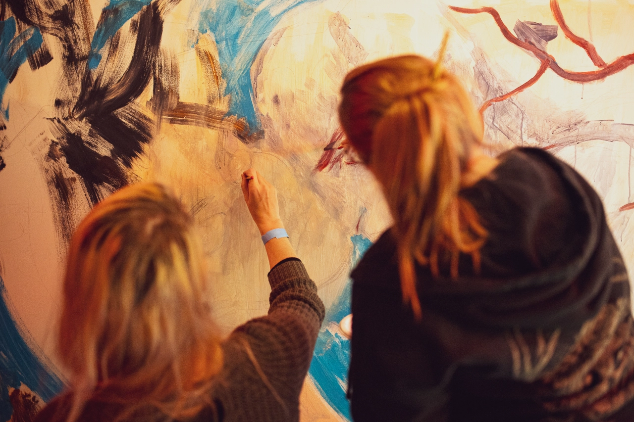 Two people with red hair painting colourful abstract designs on a large canvas wall together