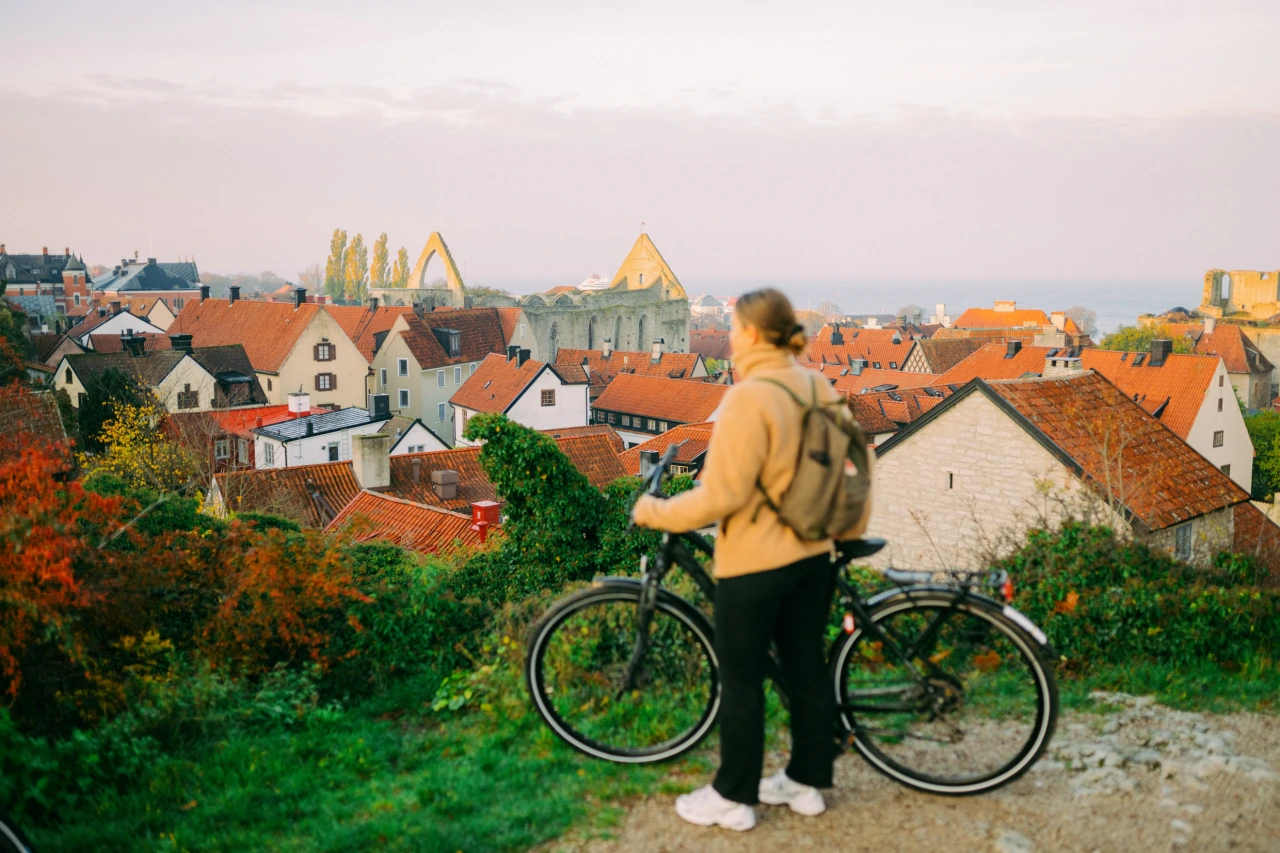 Person in yellow jacket standing with bicycle overlooking a medieval town with red-roofed houses and church spires in Visby, Sweden