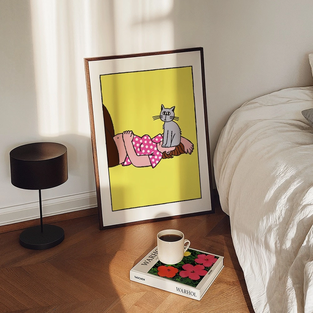 Framed illustration of a cat sitting on a person's head against a yellow background, displayed on a bedside table next to a black lamp and coffee cup