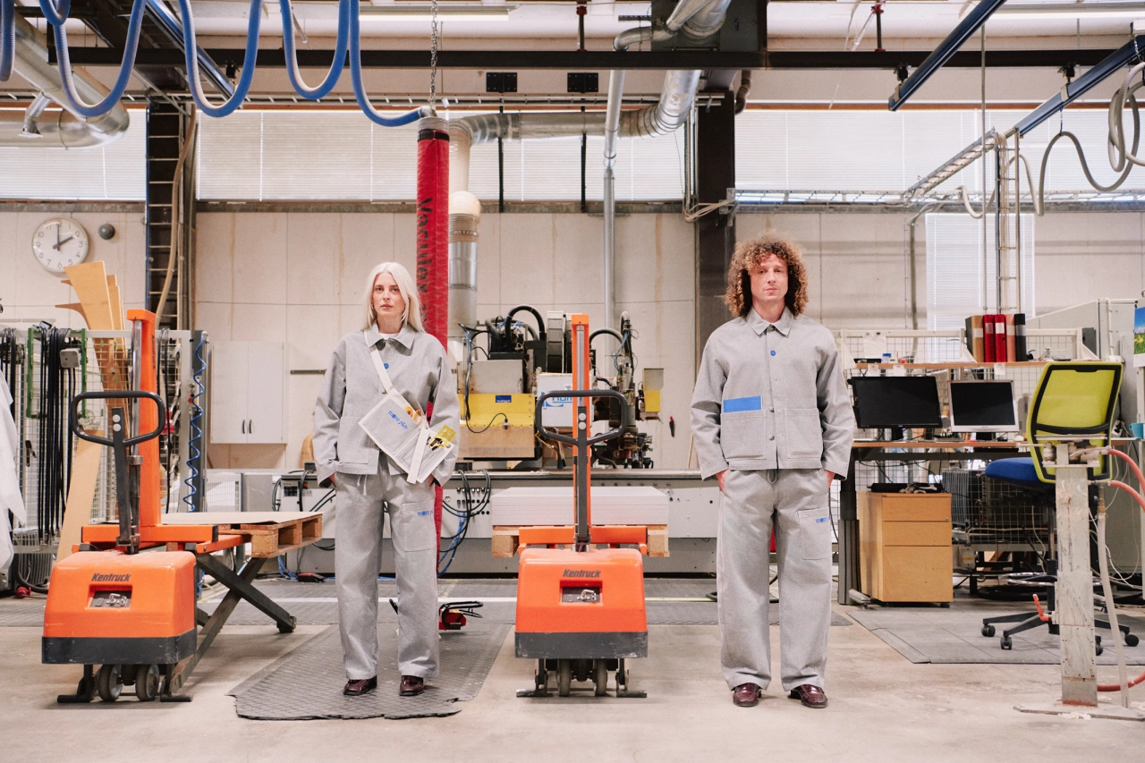Two people in grey Everyday Uniform workwear stand in Studio Playground's workshop surrounded by tools and equipment, including orange machinery.