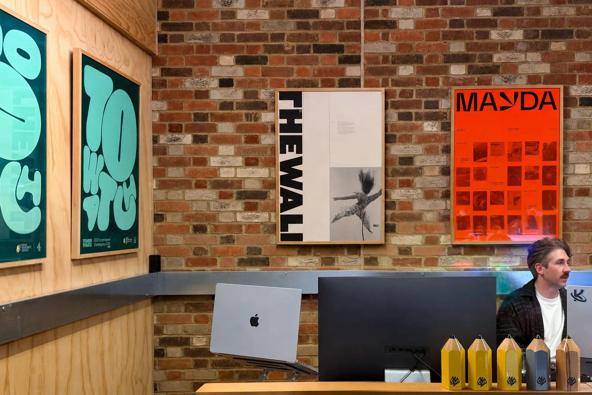 Studio Kiln's London workspace featuring green and orange branded posters on a brick wall, with laptops and design materials on a desk below