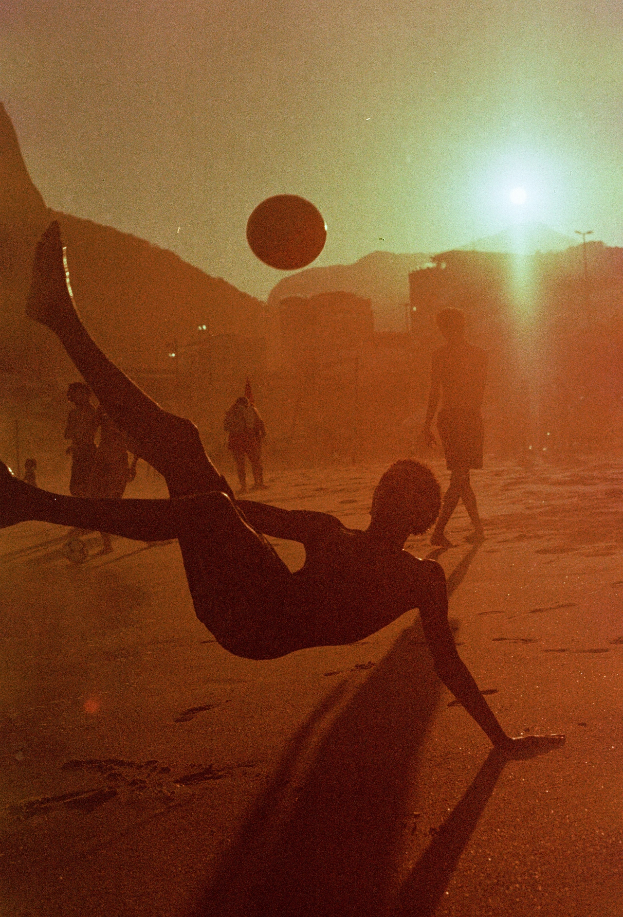 Silhouetted figure doing a backflip against a dusty orange landscape with buildings and other people in background at sunset