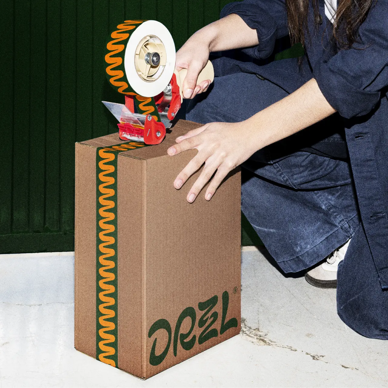 Person's hands applying orange and white tape dispenser to cardboard Drzzl olive oil box against green corrugated backdrop