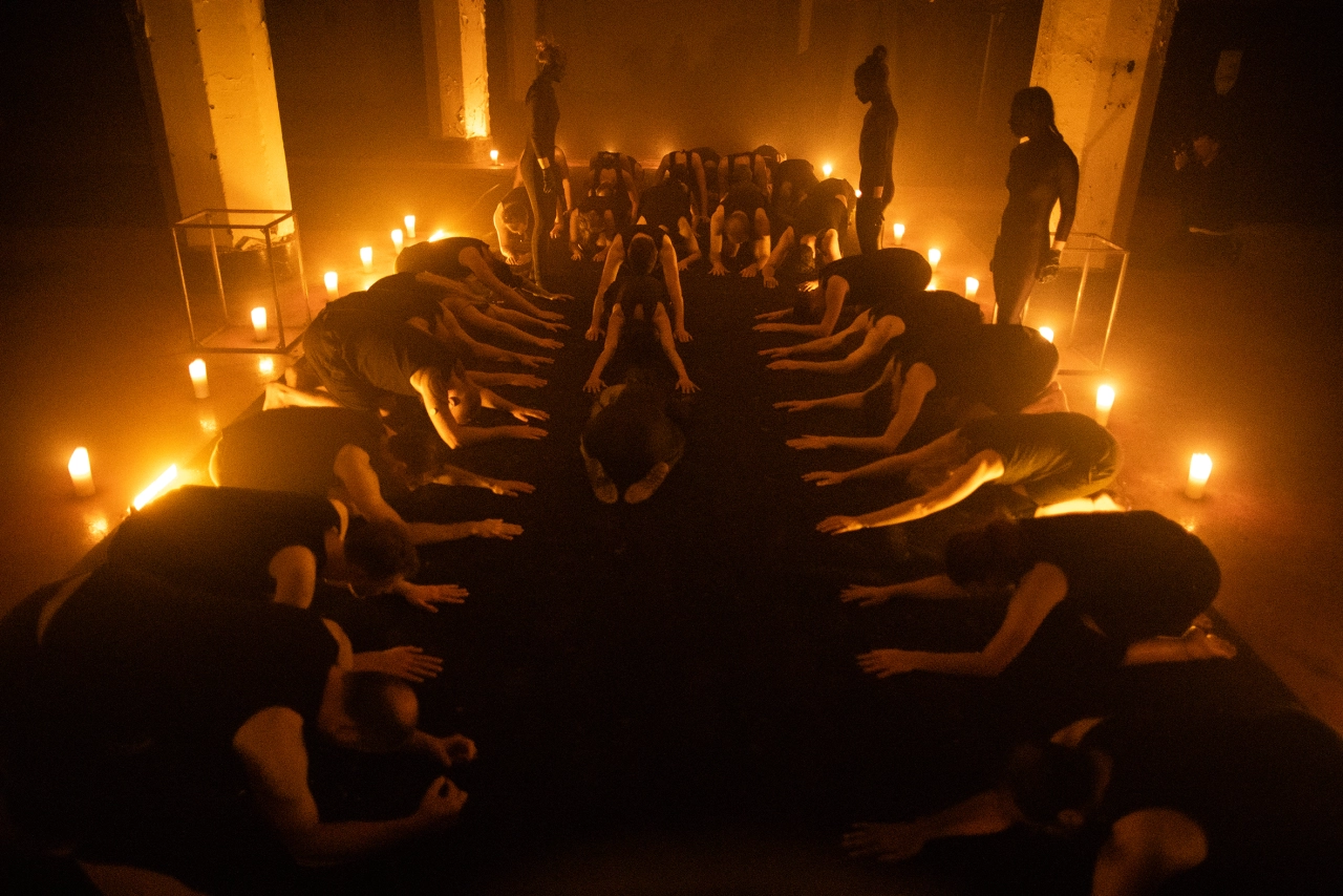 Naked participants sit in rows facing each other in a dimly lit room lined with glowing lights, creating an intense, ritualistic atmosphere