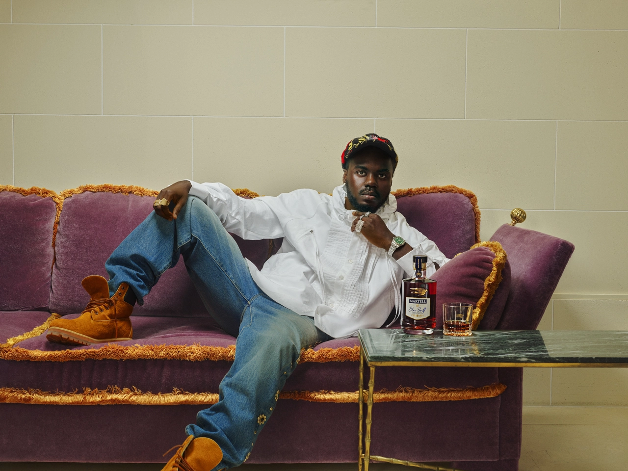 Ghanaian visual artist Prince Gyasi reclines on a purple and gold sofa holding a glass of Martell cognac against a neutral tiled wall