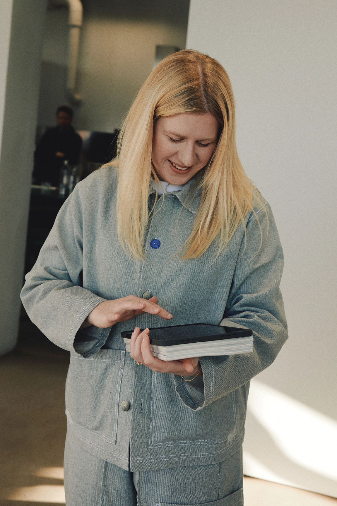 Woman with blonde hair wearing a grey shirt and matching trousers from the Everyday Uniform collection, holding a black notebook and smiling downwards