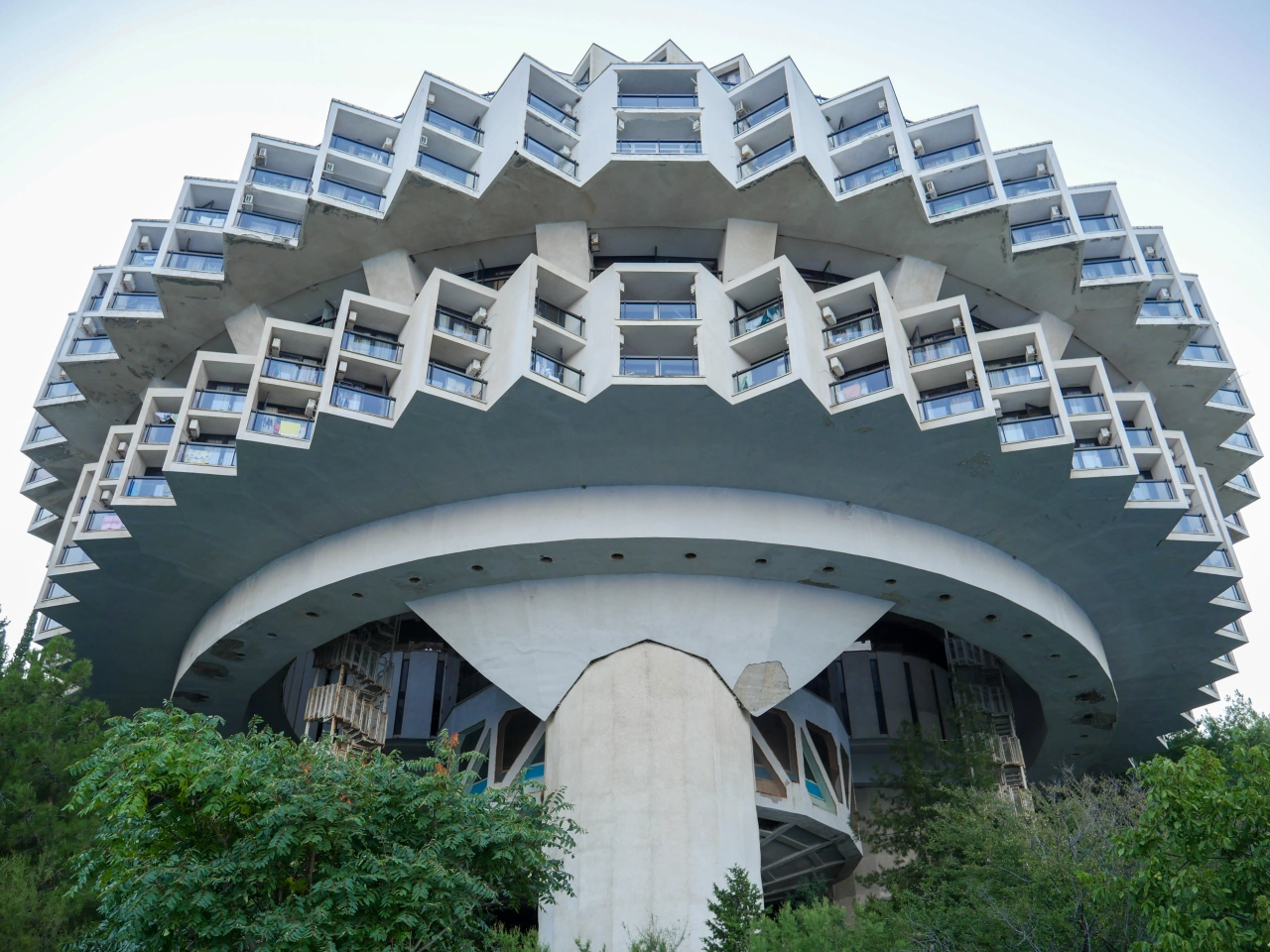 Brutalist concrete hotel tower with stacked angular balconies resembling a flying saucer, supported by a cylindrical column, surrounded by vegetation