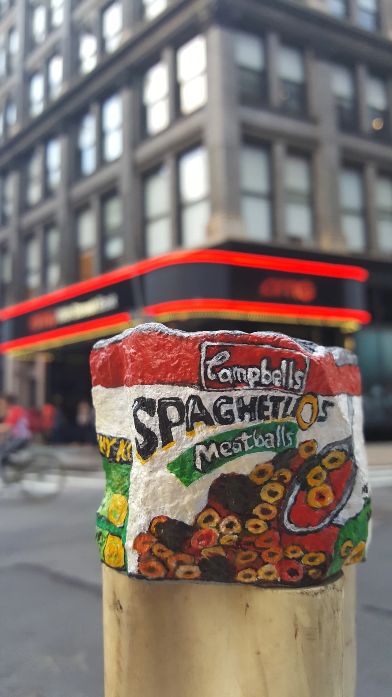 Painted rock depicting a Campbell's Spaghetti and Meatballs tin can, displayed on a urban street pole with a blurred building facade behind