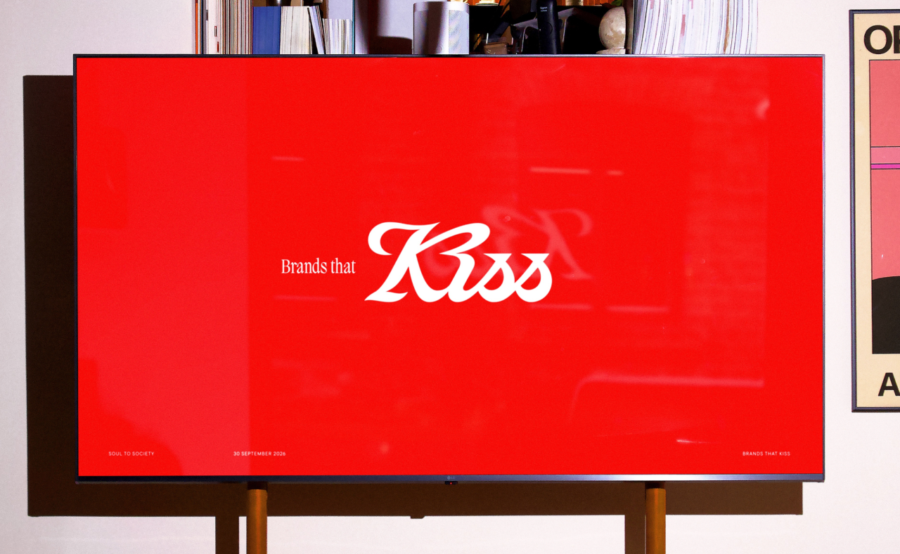 Bright red display screen showing Kiss Branding's new identity with "Brands that Kiss" tagline, mounted in an office setting with bookshelves visible behind it