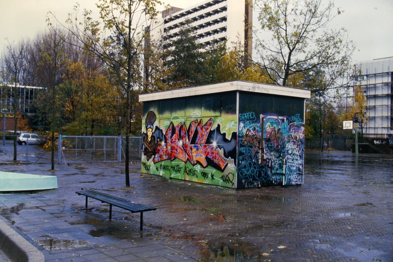 Wet urban playground with a small kiosk covered in vibrant red and blue graffiti art, surrounded by autumn trees and apartment blocks