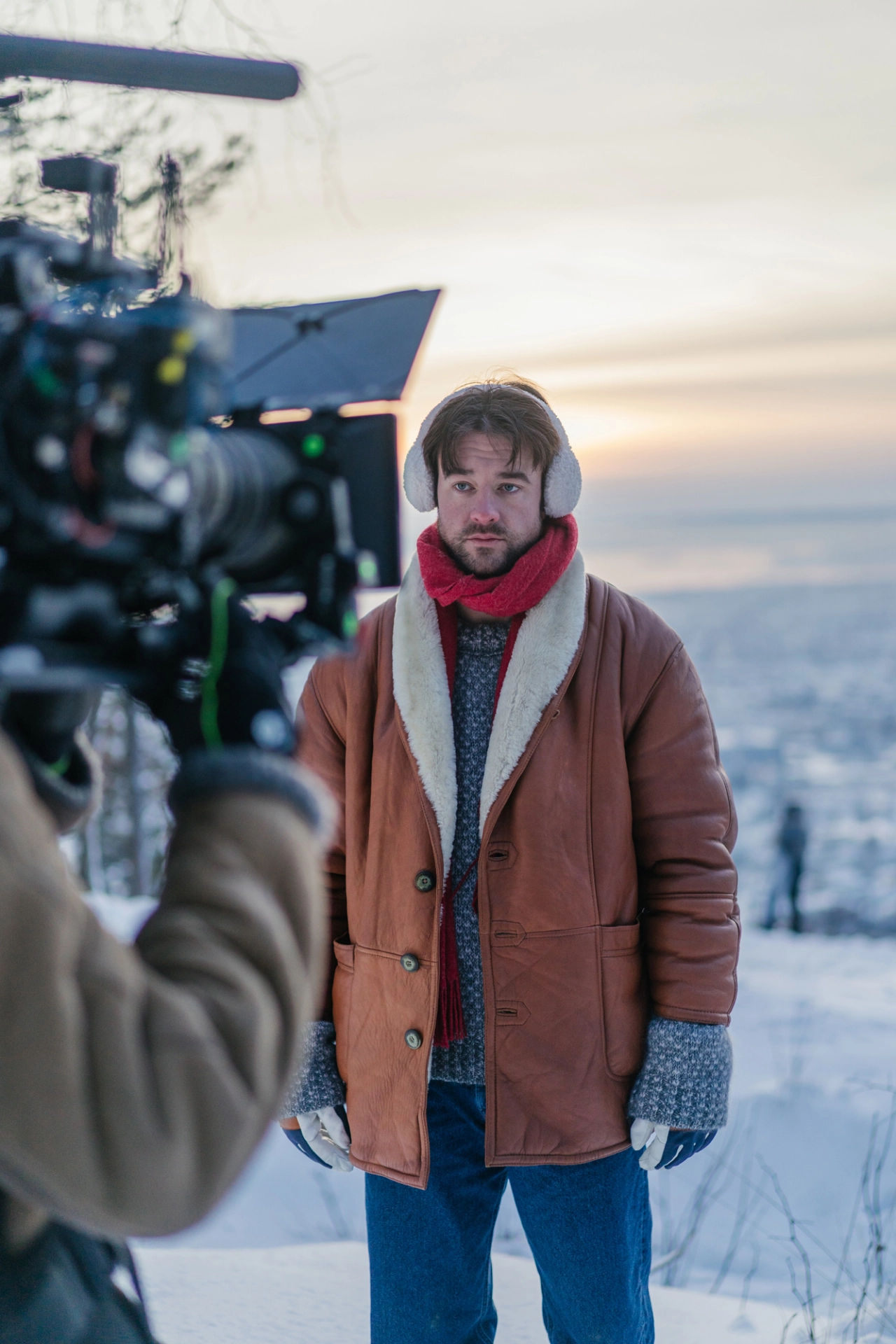 A man wearing a brown jacket, red scarf and headphones stands in front of a film camera during a snowy winter shoot at dusk