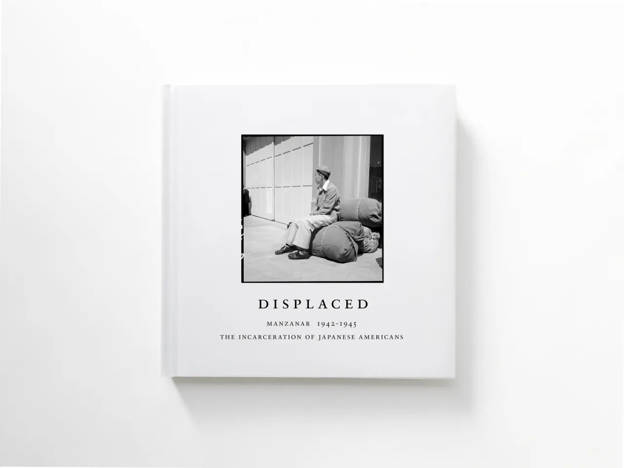 Book cover titled Displaced showing a black and white photograph of a person sitting on a cot in a sparse room, documenting Manzanar 1942-1943 and the incarceration of Japanese Americans