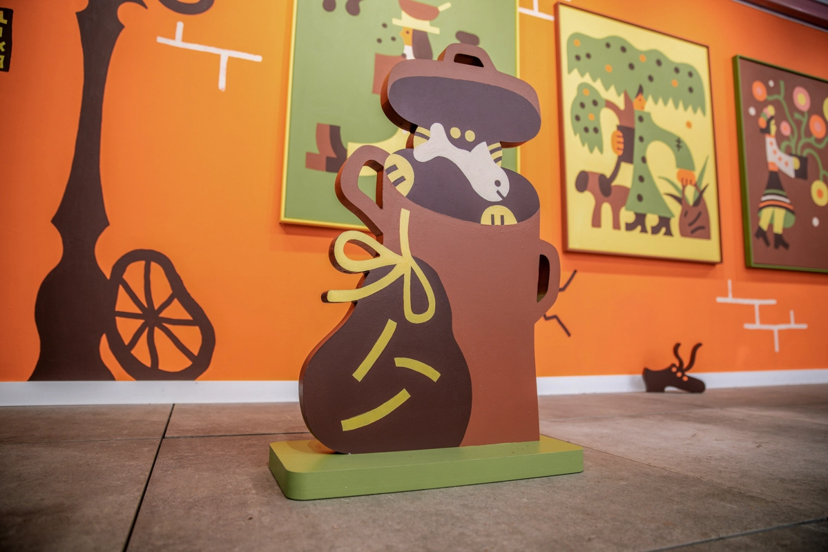Wooden sculpture of a figure wearing a hat and holding a rope, painted in brown and yellow tones, displayed on a green base against an orange gallery wall
