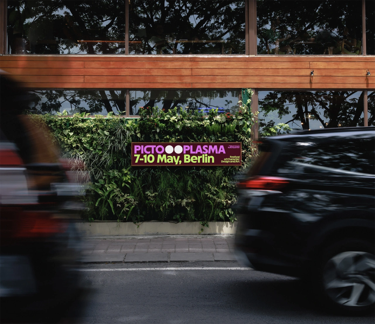 Pictoplasma festival banner with purple logotype and dates 7-10 May, Berlin displayed on a planter with green foliage, with blurred vehicles passing by on the street below