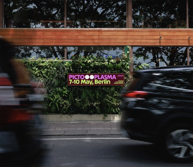 Pictoplasma festival banner with purple logotype and dates 7-10 May, Berlin displayed on a planter with green foliage, with blurred vehicles passing by on the street below