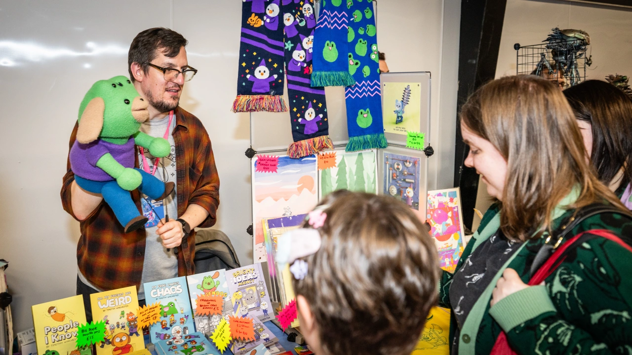 Man holding a colourful plush toy speaks to children at a Thought Bubble festival event, with comic books and artwork displayed on the table and wall behind him