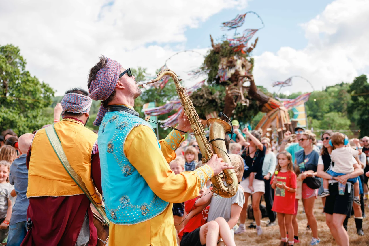 A musician wearing a colorful headwrap and yellow-and-teal outfit plays a brass saxophone outdoors in front of a crowd at a festival.