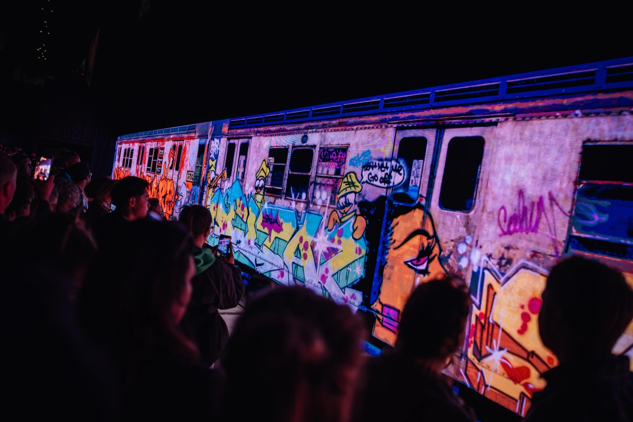A colourfully graffitied train carriage illuminated in pink and blue light, surrounded by silhouetted spectators viewing the artwork at night