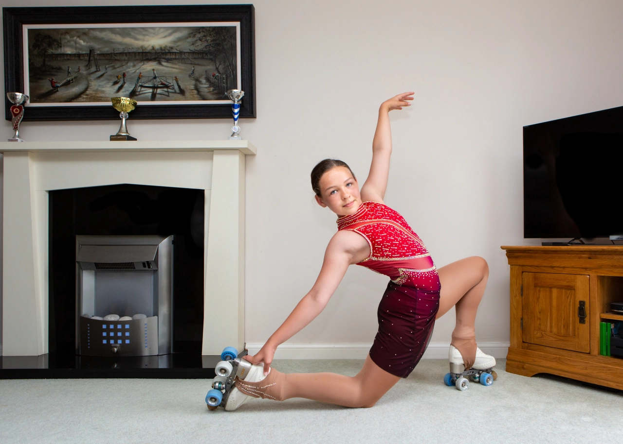 Young roller skater performing a flexible skating move on a living room floor, wearing a red sequinned costume and roller skates