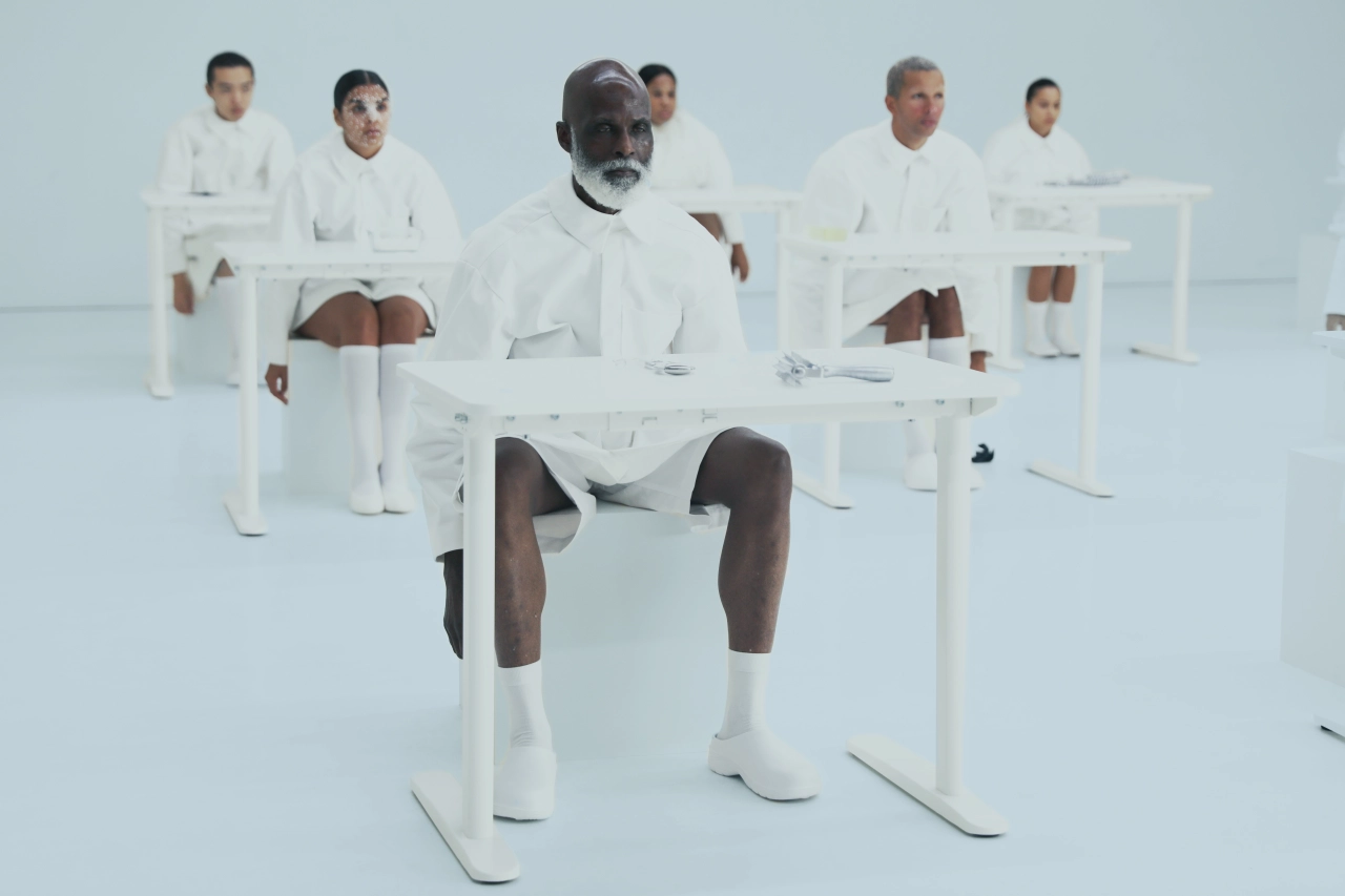 Group of people seated on white chairs wearing white clothing against a light blue background in a minimalist studio setting