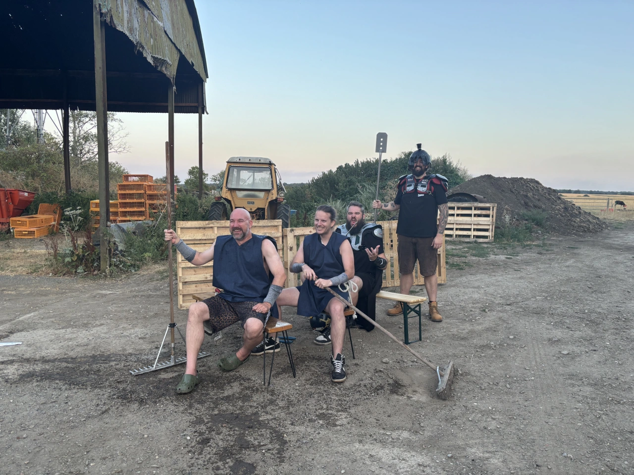 A group of filmmakers sit and stand on chairs in a rural farmyard setting with a crane, tractor and wooden pallets visible in the background