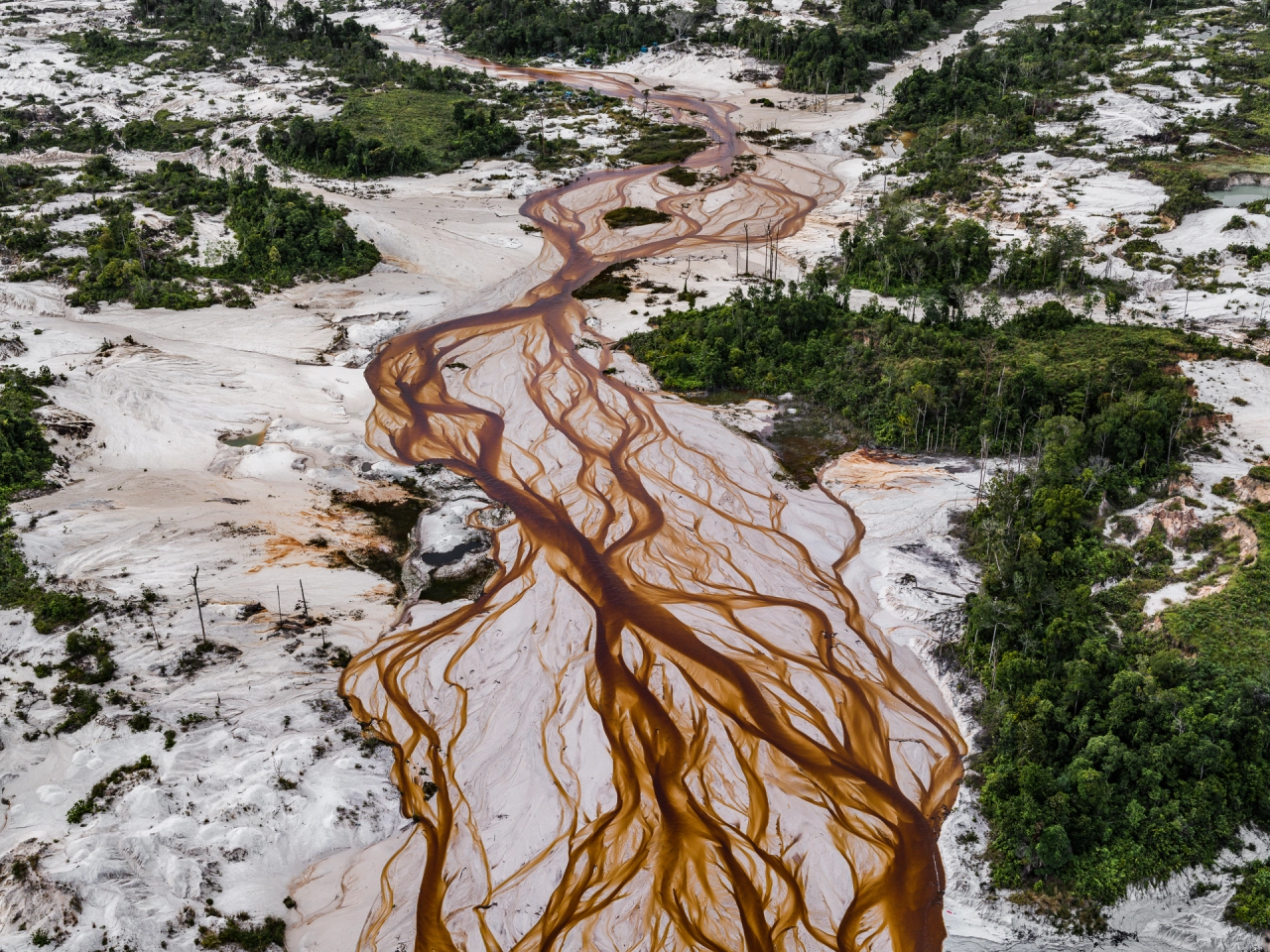 Aerial view of a geothermal hot spring with rust-coloured mineral deposits branching across white mineral-rich ground surrounded by green forest