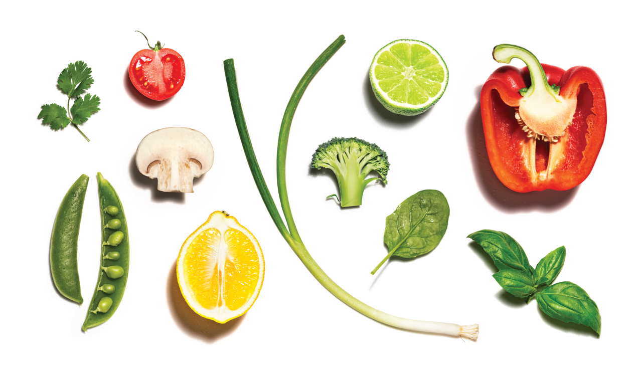 Flat lay arrangement of fresh ingredients including tomato, red pepper, lime, mushroom, peas, lemon, spinach, basil, parsley and spring onion on white background