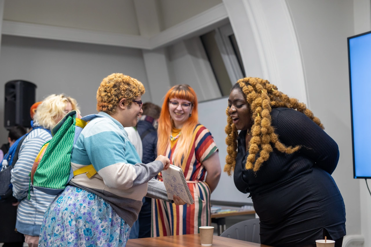 Three people smiling and chatting at an indoor event, including someone with golden curled braids wearing a black top and glasses, and another person with orange fringe and a striped top