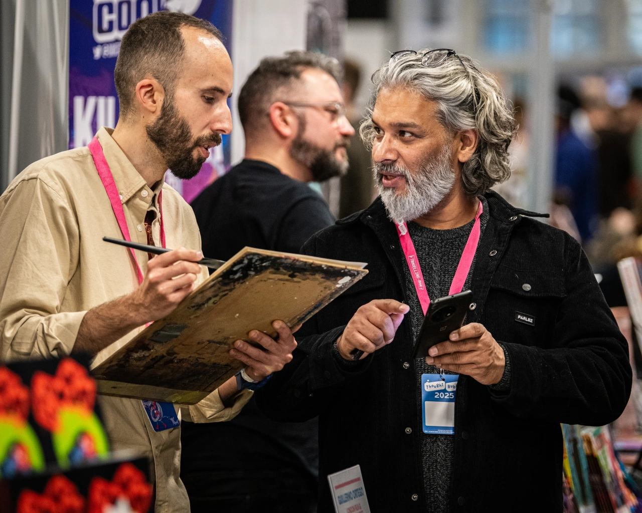 Two men wearing conference badges examine an artwork together at Thought Bubble festival, with another attendee visible in the background