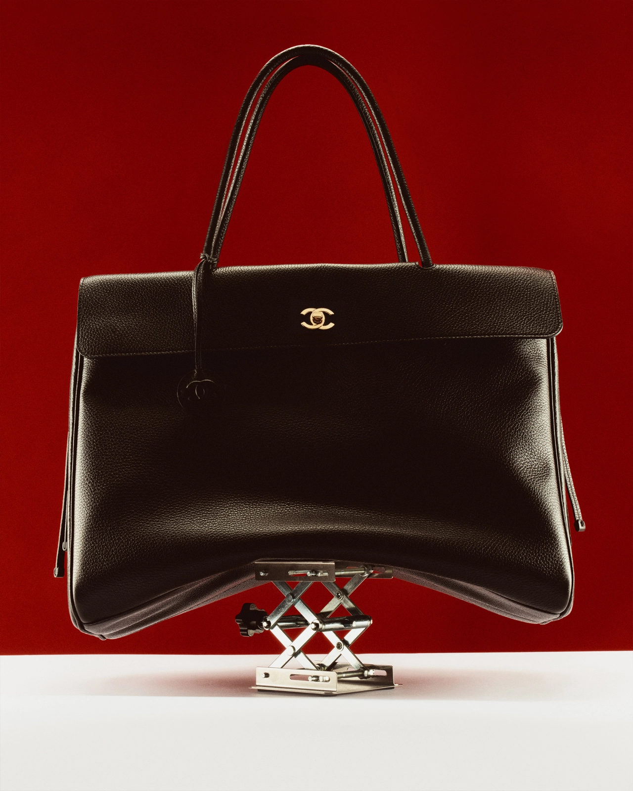 Black leather Chanel handbag with gold logo perched on a white scissor jack against a deep red background