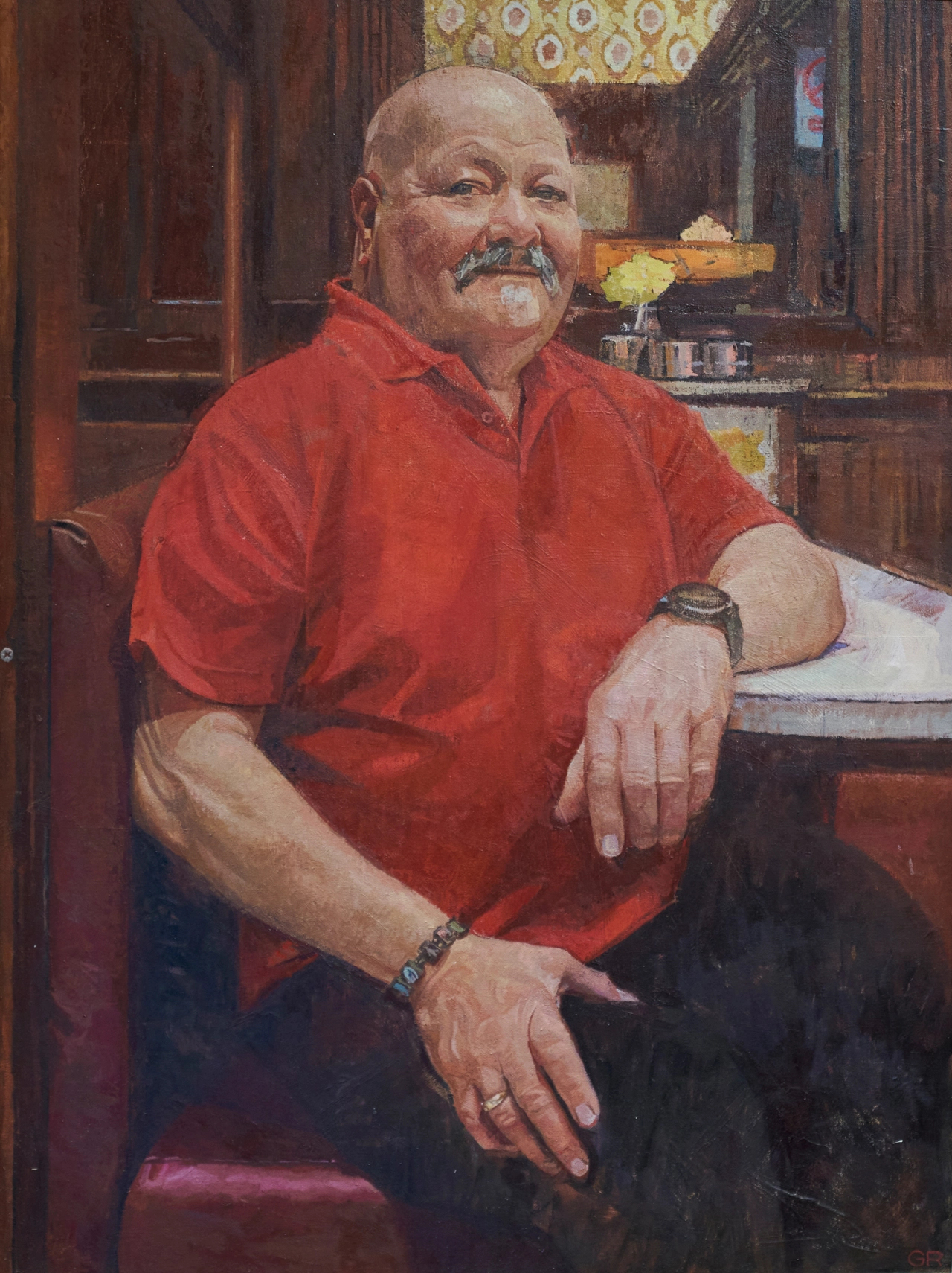 Bald man with a moustache wearing a red shirt, seated in a wood-panelled interior with a yellow flower visible behind him