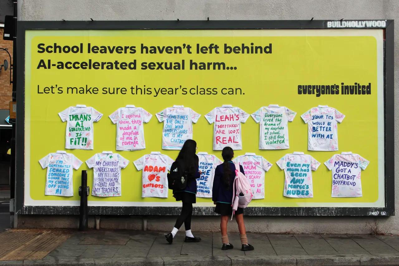 Two young people view a billboard displaying white school leavers' shirts with handwritten testimonies about AI-enabled sexual harm, part of the Everyone's Invited campaign