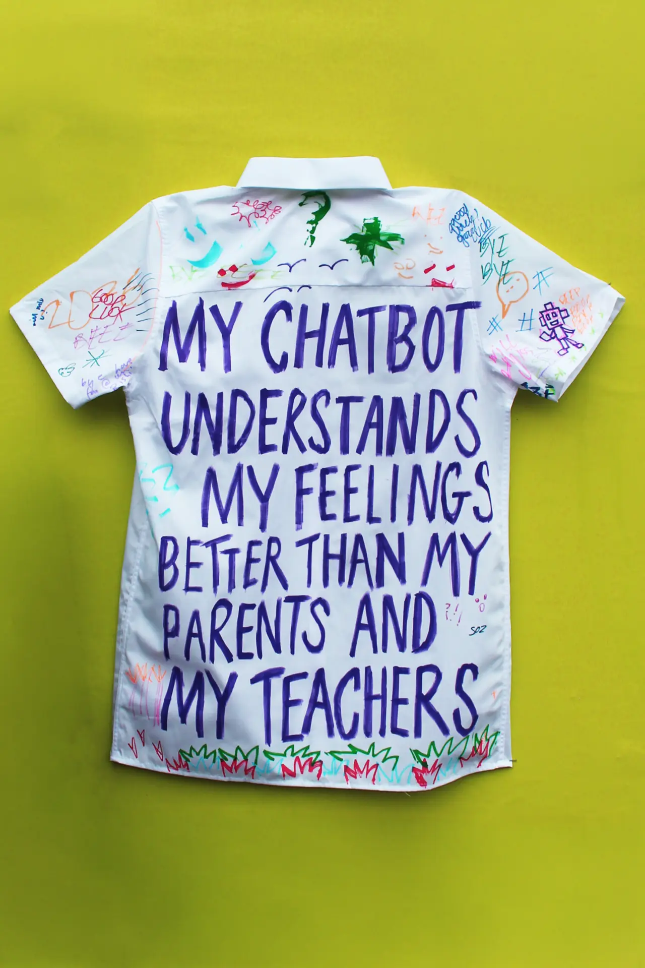 White school leavers' shirt with blue text reading "My chatbot understands my feelings better than my parents and my teachers" and colourful doodles, against a bright yellow background