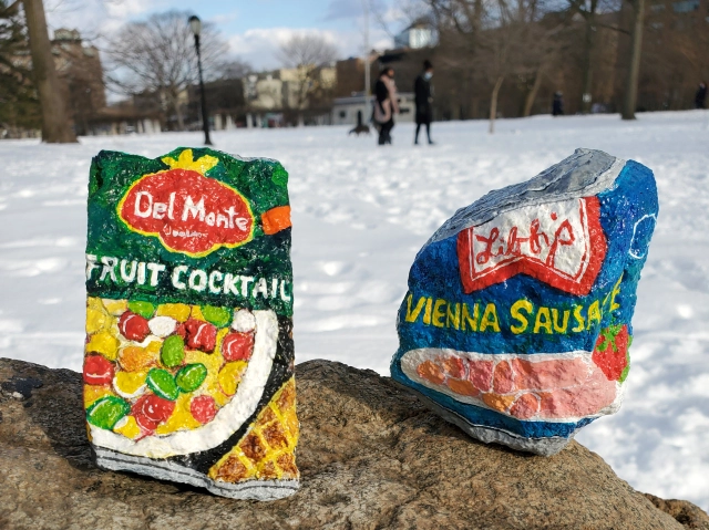 Two painted rocks on a stone surface in a snowy park: one depicting a Del Monte fruit cocktail tin and the other a blue Libra Vienna sausage tin