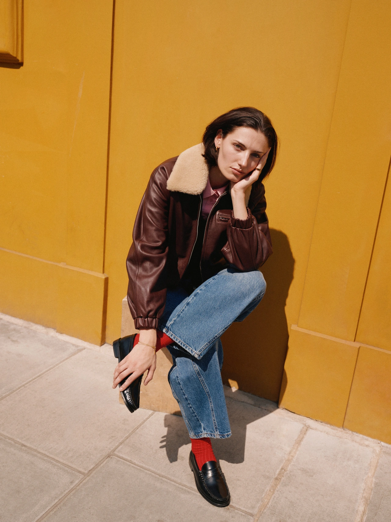 Woman in burgundy leather jacket with sheepskin collar and blue jeans crouching against a bright yellow wall