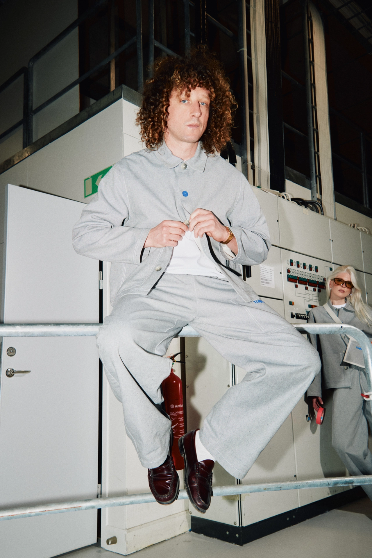 Person wearing light grey workwear uniform consisting of shirt and trousers, designed by Oxox Club for Studio Playground, seated on industrial equipment in a studio setting